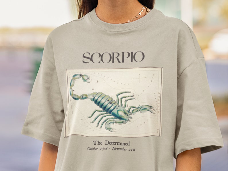 Op de afbeelding: Een lichtgrijs T-shirt met een vintage illustratie van het sterrenbeeld Schorpioen en de tekst "SCORPIO" en "The Determined October 23rd - November 21st".