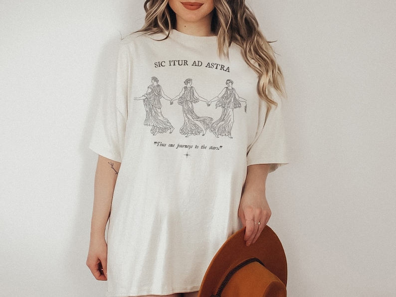 May include: A white t-shirt with a graphic of three women holding hands and the text "Sic Itur Ad Astra" and "Thus one journeys to the stars".