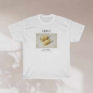 May include: White t-shirt featuring a Libra zodiac design. The graphic includes the word "LIBRA" above an illustration of the Libra symbol, with the text "The Charmer" and the dates "September 23rd - October 22nd" below the illustration. The shirt is made of a soft material.