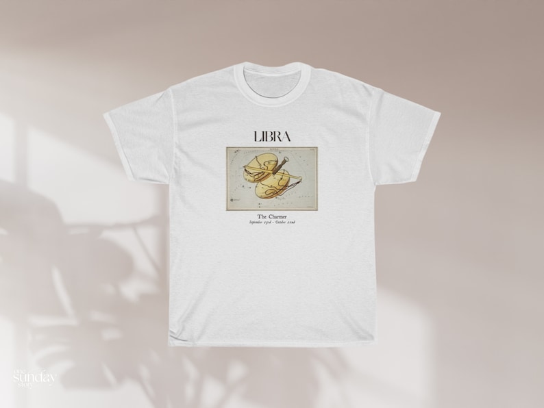 May include: White t-shirt featuring a Libra zodiac design. The graphic includes the word "LIBRA" above an illustration of the Libra symbol, with the text "The Charmer" and the dates "September 23rd - October 22nd" below the illustration. The shirt is made of a soft material.
