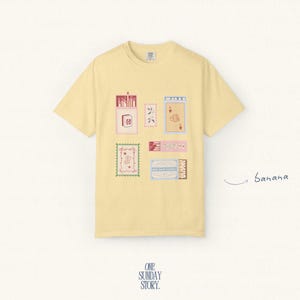 May include: A pale yellow short-sleeve t-shirt featuring a graphic print of various rectangular illustrations including matchboxes, playing cards, and decorative labels. The word banana is written in cursive script to the right of the shirt.