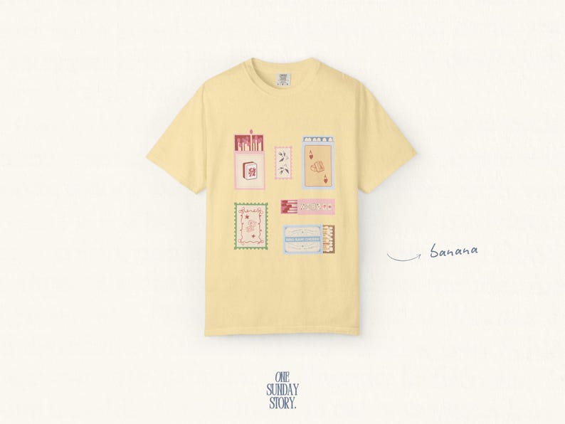 May include: A pale yellow short-sleeve t-shirt featuring a graphic print of various rectangular illustrations including matchboxes, playing cards, and decorative labels. The word banana is written in cursive script to the right of the shirt.