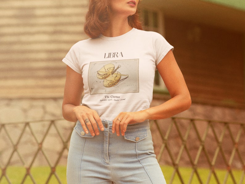May include: White t-shirt featuring a Libra zodiac design with a violin illustration. The text "LIBRA" is above the graphic, and "The Charmer September 23rd - October 22nd" is below. The shirt is paired with light blue jeans.