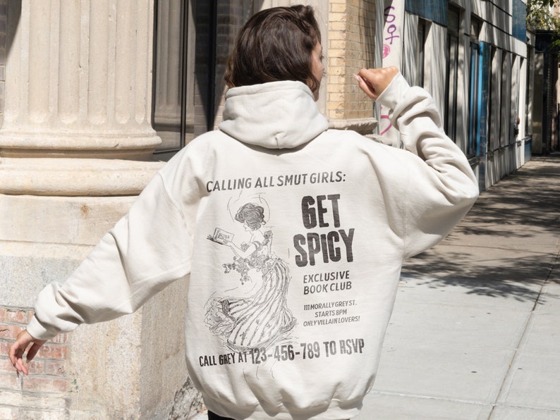 May include: A cream-colored hoodie with a black graphic print on the back. The graphic features a woman reading a book with the text "Get Spicy" and "Exclusive Book Club".