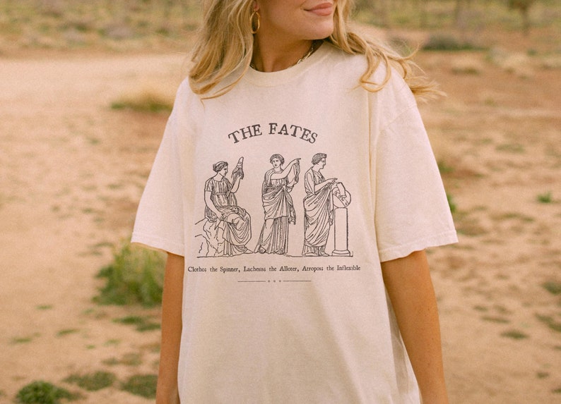 May include: A white t-shirt with a graphic print of three women representing the Fates. The text "THE FATES" is printed above the graphic. Below the graphic, the text reads "Clotho the Spinner, Lachesis the Allotter, Atropos the Inevitable."