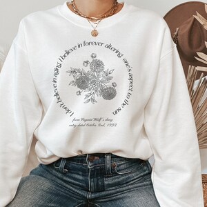 May include: White sweatshirt with a black floral design and the text "I don't believe in aging. I believe in forever altering one's aspect to the sun. from Virginia Woolf's diary entry dated October 2nd, 1932"