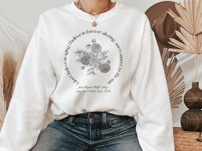 May include: White sweatshirt with a black floral design and the text "I don't believe in aging. I believe in forever altering one's aspect to the sun. from Virginia Woolf's diary entry dated October 2nd, 1932"