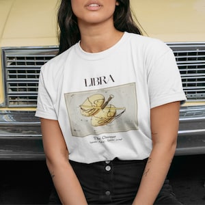 May include: White t-shirt featuring the word "LIBRA" above a vintage-style illustration of a scale and the words "The Charmer". The shirt is a classic crew neck style and is made of a soft, comfortable material.