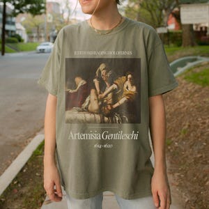 May include: A green t-shirt with a vintage-style print of a painting titled "Judith Beheading Holofernes" by Artemisia Gentileschi (1614-1620).