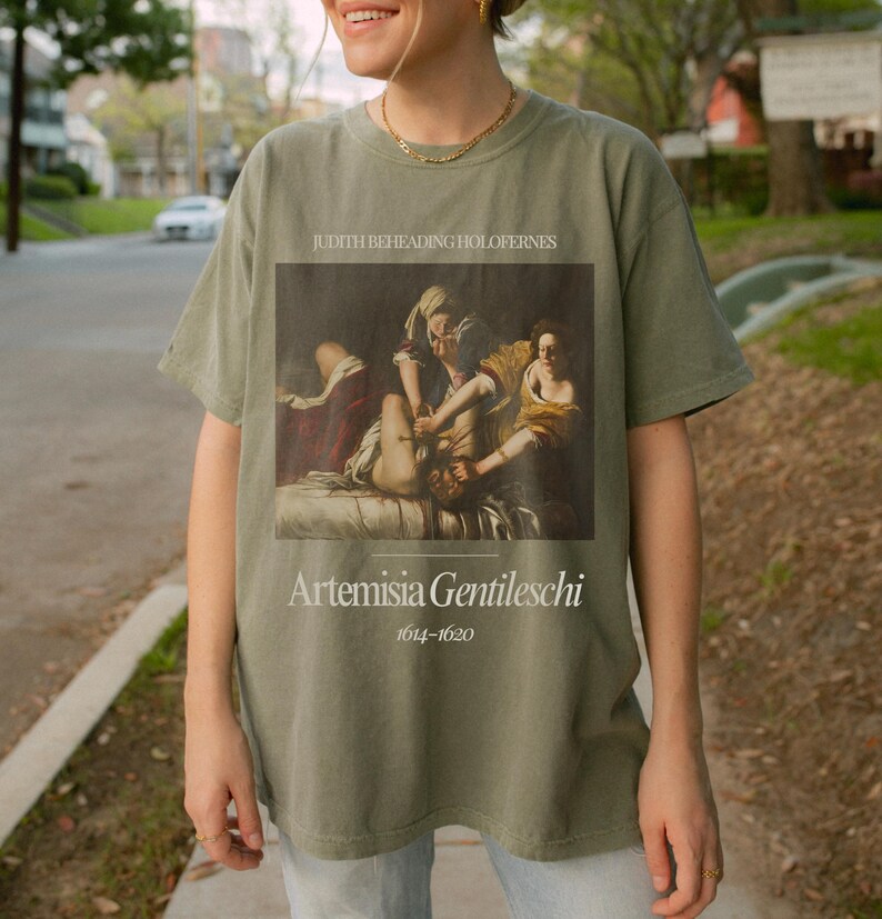 May include: A green t-shirt with a vintage-style print of a painting titled "Judith Beheading Holofernes" by Artemisia Gentileschi (1614-1620).