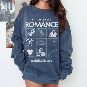 Puede incluir: Una sudadera azul marino con el texto "HOT GIRLS READ ROMANCE" y una cuadr&iacute;cula de nueve ilustraciones blancas. Las ilustraciones incluyen un coraz&oacute;n, un libro, un lazo, una pluma, una flecha, una flor y tres figuras. El texto "A BOOK CLUB BY BOOKED READER GIRLS" est&aacute; impreso debajo de la cuadr&iacute;cula.