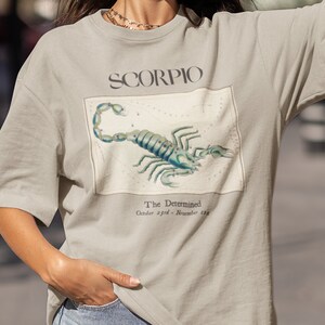 Op de afbeelding: Een licht beige t-shirt met een grafische print van een schorpioen en de tekst "SCORPIO The Determined October 23rd - November 21st".
