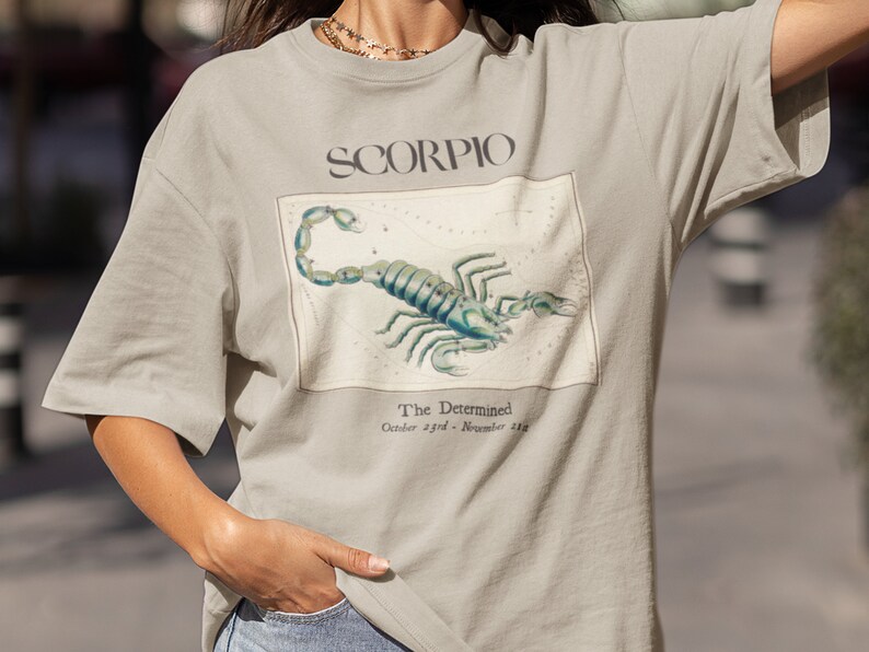 Op de afbeelding: Een licht beige t-shirt met een grafische print van een schorpioen en de tekst "SCORPIO The Determined October 23rd - November 21st".