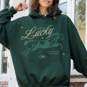 May include: Dark green hoodie with a kangaroo pocket and a drawstring hood. The front features a graphic of a running horse with the word "Lucky" in a script font. Text below reads "Year of the fire horse" and "A year ruled by strength, motion, and fortune."