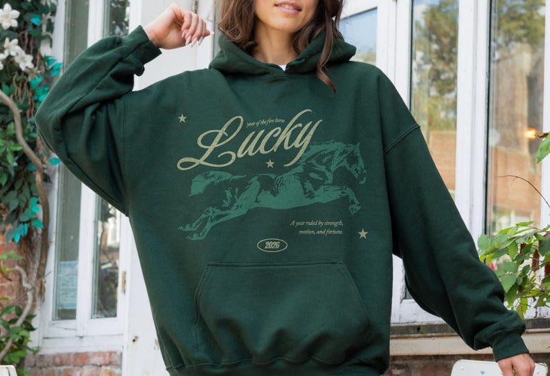 May include: Dark green hoodie with a kangaroo pocket and a drawstring hood. The front features a graphic of a running horse with the word "Lucky" in a script font. Text below reads "Year of the fire horse" and "A year ruled by strength, motion, and fortune."