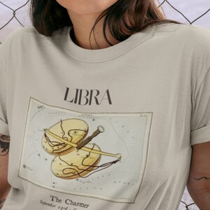 May include: Beige t-shirt featuring the word "LIBRA" above a vintage-style illustration of the Libra zodiac symbol. The illustration includes a scale design with the text "The Charmer" and "September 23rd - October 22nd".