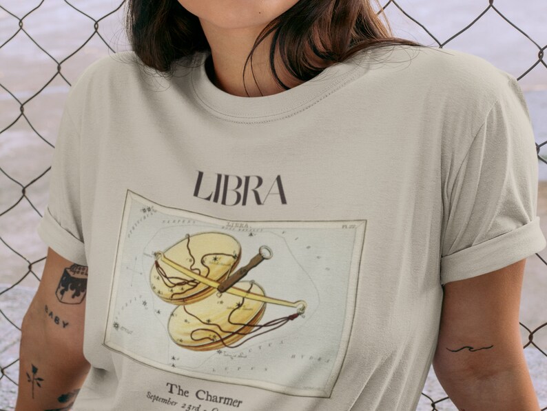 May include: Beige t-shirt featuring the word "LIBRA" above a vintage-style illustration of the Libra zodiac symbol. The illustration includes a scale design with the text "The Charmer" and "September 23rd - October 22nd".