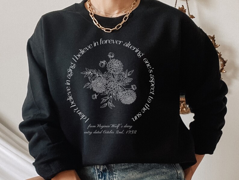 May include: Black sweatshirt with a white graphic print of flowers and text that reads "don't believe in aging I believe in forever altering one's aspect to the sun" and "from Virginia Woolf's diary entry dated October 2nd, 1932".