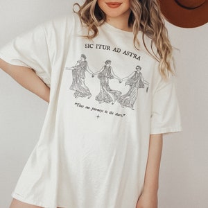 May include: A white oversized t-shirt with a black graphic print of three women holding hands. The text "Sic Itur Ad Astra" is printed above the graphic and the text "my thus one journeys to the stars." is printed below the graphic.