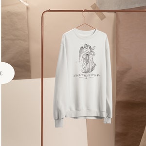 May include: White sweatshirt with a black and white illustration of two angels embracing. The text "AMOR VINCIT OMNIA" is printed below the illustration.