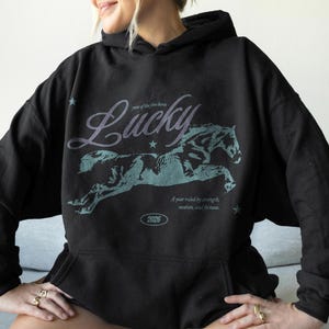May include: Black hoodie featuring a running horse graphic and the word "Lucky". The text "A year ruled by strength, wisdom, and fortune" is below the horse. The hoodie has a drawstring hood and a kangaroo pocket.
