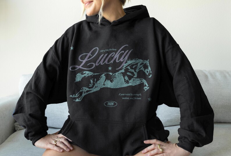 May include: Black hoodie featuring a running horse graphic and the word "Lucky". The text "A year ruled by strength, wisdom, and fortune" is below the horse. The hoodie has a drawstring hood and a kangaroo pocket.