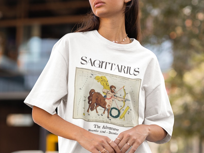 May include: White t-shirt with a vintage illustration of the Sagittarius zodiac sign. The illustration is in brown, yellow, and black. The text "SAGITTARIUS" is printed in black above the illustration. The text "The Adventurer" is printed in black below the illustration. The text "November 22nd - December 21st" is printed in black below the text "The Adventurer."