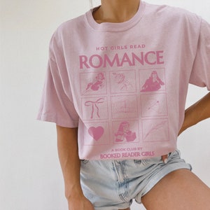May include: A pink t-shirt with the text "HOT GIRLS READ ROMANCE" and a grid of illustrations. The text "A BOOK CLUB BY BOOKED READER GIRLS" is at the bottom of the shirt.