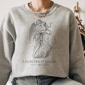 May include: A gray sweatshirt with a black and white illustration of a couple embracing. The text "AMOR VINCIT OMNIA" is printed below the illustration.