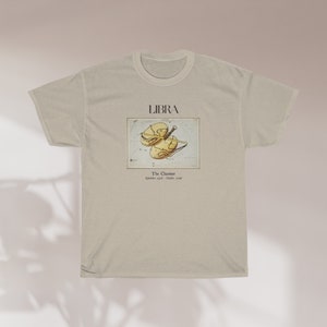 May include: A beige t-shirt featuring the Libra zodiac sign. The design includes the word "LIBRA" above an illustration of the scales of justice. Below the illustration is the text "The Chariot" and the dates "September 23rd - October 22nd."