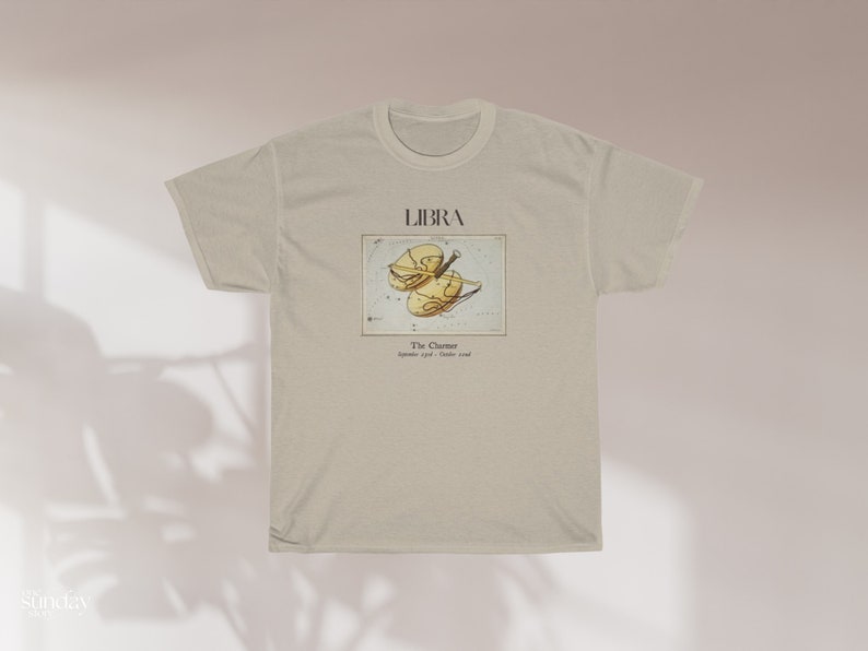May include: A beige t-shirt featuring the Libra zodiac sign. The design includes the word "LIBRA" above an illustration of the scales of justice. Below the illustration is the text "The Chariot" and the dates "September 23rd - October 22nd."