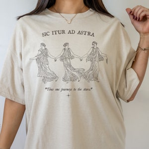 May include: A beige t-shirt with a black graphic print of three women holding hands. The text "Sic Itur Ad Astra" and "Thus one journeys to the stars." is printed on the shirt.