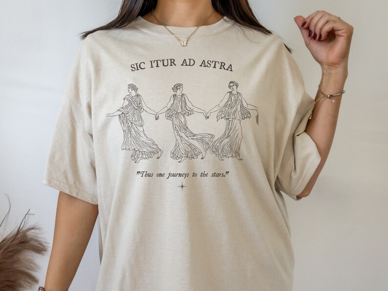 May include: A beige t-shirt with a black graphic print of three women holding hands. The text "Sic Itur Ad Astra" and "Thus one journeys to the stars." is printed on the shirt.