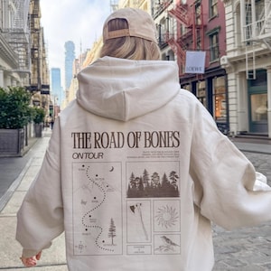 May include: Cream-colored hoodie with the text "THE ROAD OF BONES ON TOUR" in brown. The back of the hoodie features a map, forest, axe, sun, and bird illustrations. The hoodie is worn outdoors.