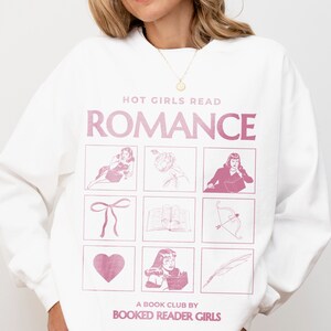 Puede incluir: Sudadera blanca con un gr&aacute;fico rosa que dice "Hot Girls Read Romance" y presenta una cuadr&iacute;cula de nueve im&aacute;genes que incluyen un coraz&oacute;n, un lazo, un libro, una flecha, una pluma y una mujer. El texto en la parte inferior dice "A Book Club by Booked Reader Girls."