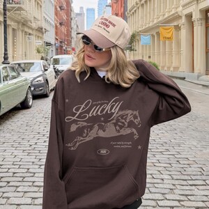May include: A brown hoodie featuring a horse graphic and the word "Lucky". The hoodie also displays the text "Year of the fire horse" and "A year ruled by strength, motion, and fortune". The person is wearing a beige cap.