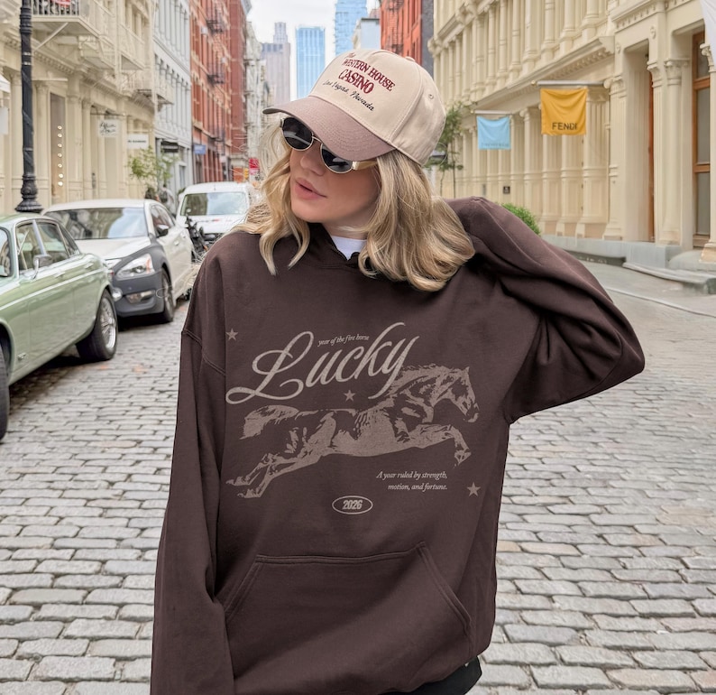 May include: A brown hoodie featuring a horse graphic and the word "Lucky". The hoodie also displays the text "Year of the fire horse" and "A year ruled by strength, motion, and fortune". The person is wearing a beige cap.
