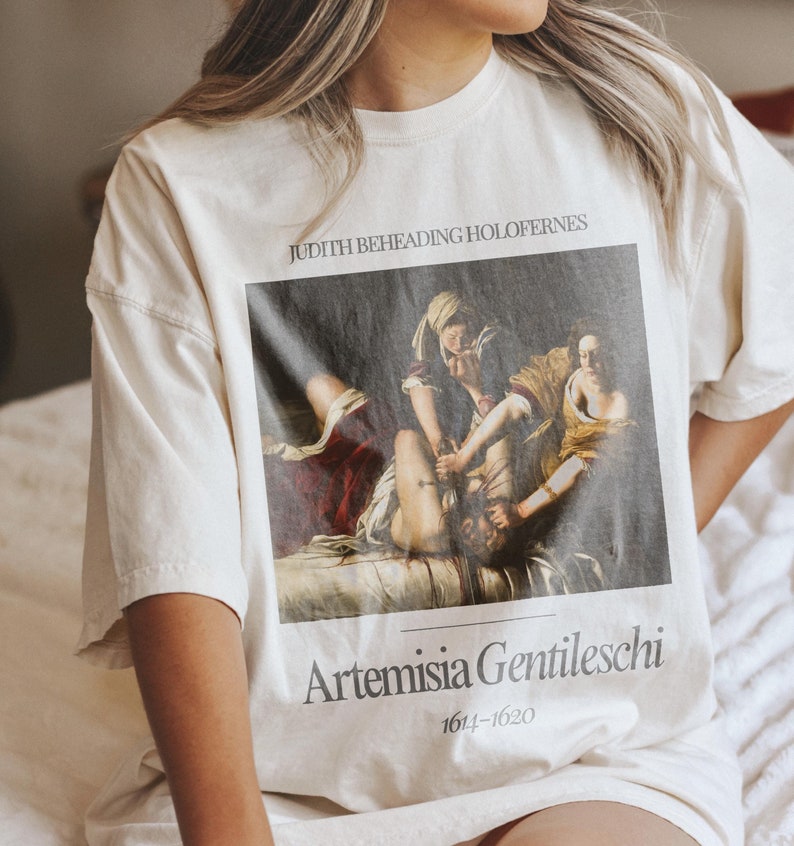 May include: A white t-shirt with a painting of Judith Beheading Holofernes by Artemisia Gentileschi. The painting is in a black frame and the text "Judith Beheading Holofernes" and "Artemisia Gentileschi 1614-1620" is printed below the painting.