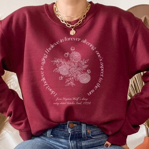 May include: Burgundy sweatshirt with a white floral design and a quote from Virginia Woolf's diary: "I don't believe in aging. I believe in forever altering one's aspect to the sun." The quote is centered on the sweatshirt and surrounded by a circle of white flowers.