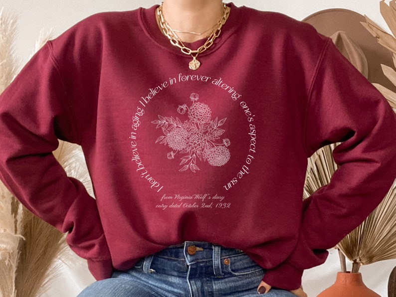 May include: Burgundy sweatshirt with a white floral design and a quote from Virginia Woolf's diary: "I don't believe in aging. I believe in forever altering one's aspect to the sun." The quote is centered on the sweatshirt and surrounded by a circle of white flowers.