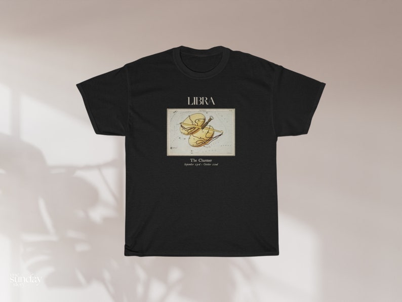 May include: Black t-shirt featuring the word "LIBRA" above a vintage-style illustration of the Libra zodiac symbol. The graphic includes the text "The Charmer" and the dates "September 23rd - October 22nd".