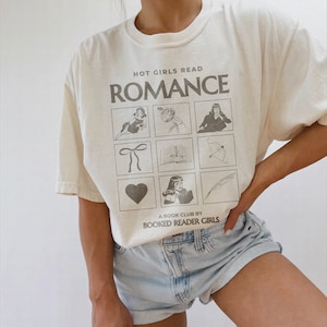 May include: A white t-shirt with the text "HOT GIRLS READ ROMANCE" and a grid of nine illustrations. The illustrations include a woman reading a book, a bow, a book, an arrow, a heart, a woman with a book, and a feather. The text "A BOOK CLUB BY BOOKED READER GIRLS" is printed below the grid.