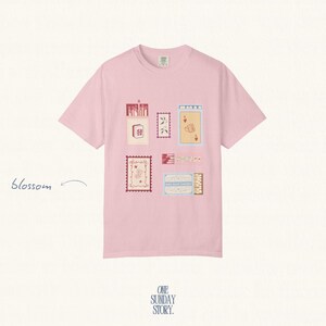 May include: A light pink short-sleeve t-shirt featuring a graphic print of various rectangular postage stamp-style illustrations in shades of red, cream, and blue. The word blossom is printed in cursive to the left of the shirt design.