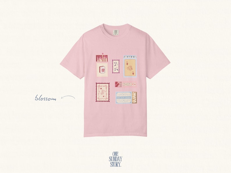 May include: A light pink short-sleeve t-shirt featuring a graphic print of various rectangular postage stamp-style illustrations in shades of red, cream, and blue. The word blossom is printed in cursive to the left of the shirt design.