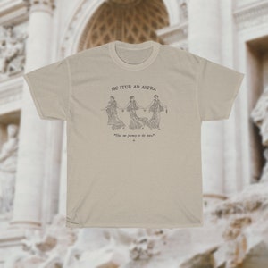 May include: A beige t-shirt with a black and white illustration of three women holding hands. The text "SIC ITUR AD ASTRA" is above the illustration and the text "The one journey is the stars" is below the illustration.