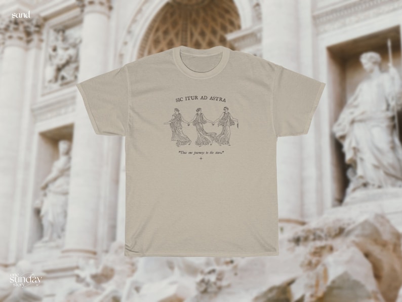 May include: A beige t-shirt with a black and white illustration of three women holding hands. The text "SIC ITUR AD ASTRA" is above the illustration and the text "The one journey is the stars" is below the illustration.