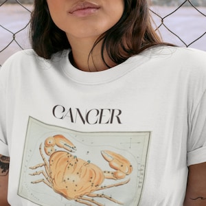 May include: White t-shirt with the word "Cancer" printed in black and a vintage illustration of the Cancer zodiac sign, a crab.