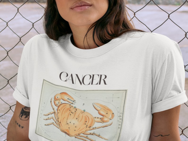 May include: White t-shirt with the word "Cancer" printed in black and a vintage illustration of the Cancer zodiac sign, a crab.