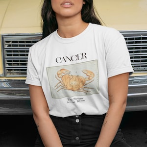 May include: White t-shirt with a graphic of the Cancer zodiac sign, a crab, and the text "Cancer The Nurturer You Feel, I See".