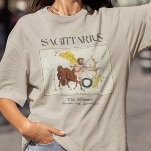 May include: A beige t-shirt with a vintage illustration of the Sagittarius constellation and the text "SAGITTARIUS The Adventurer November 22nd - December 21st".